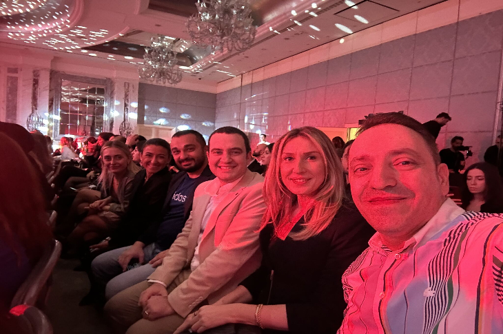 Sıra sıra yan yana oturmuş, şık giyimli altı kişi bir salonda etkinlik izliyor. Loş, kırmızımsı ışık ortamı sıcak bir hava yaratmış. Herkes kameraya doğru gülümseyerek poz veriyor; arkada da kalabalık bir izleyici kitlesi görünüyor.