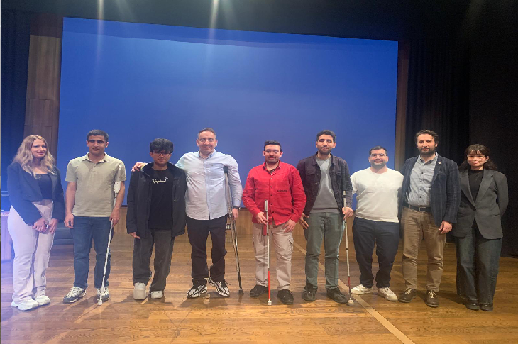 Sahnedeki fotoğrafta, dokuz kişilik bir grup yan yana dizilmiş hâlde poz veriyor. Arka planda büyük, mavi bir sunum perdesi var; ortam bir konferans salonu ya da sahne hissi veriyor. Grubun içinde hem kadın hem erkekler bulunuyor; bazı kişiler beyaz bastonlarıyla görme engelli olduklarını gösteriyor. Orta bölümde duran dört erkek birbirine yakın ve destekleyici bir duruş sergiliyor. Herkes kameraya dönük, sıcak ve samimi bir ifade ile gülümsüyor. Genel atmosfer: dayanışma, eğitim, birlikte üretme ruhu.