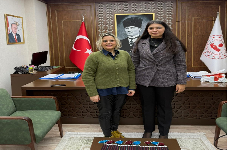 Görselde bir makam odasında yan yana duran iki kadın görülüyor. Arkalarında Türkiye ve kurum bayrakları ile Atatürk portresi yer alıyor. Odada ahşap bir masa, üzerinde dosyalar ve belgeler, önde de yeşil koltuklar bulunuyor. Kadınlar gülümseyerek poz veriyor; ortam resmi ama sıcak bir ziyaret havası taşıyor.