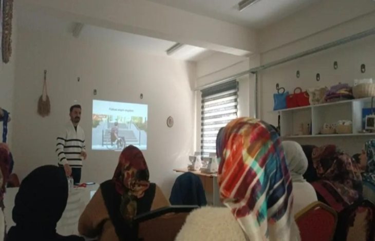 Bir sınıf ortamında, başörtülü kadınlardan oluşan bir grup sandalyelerde oturmuş eğitmene doğru bakıyor. Eğitmen, projeksiyonla yansıtılan “Fiziksel erişim engelleri” başlıklı bir görsel eşliğinde sunum yapıyor. Odada raflarda çantalar ve el işi ürünler de görülüyor.