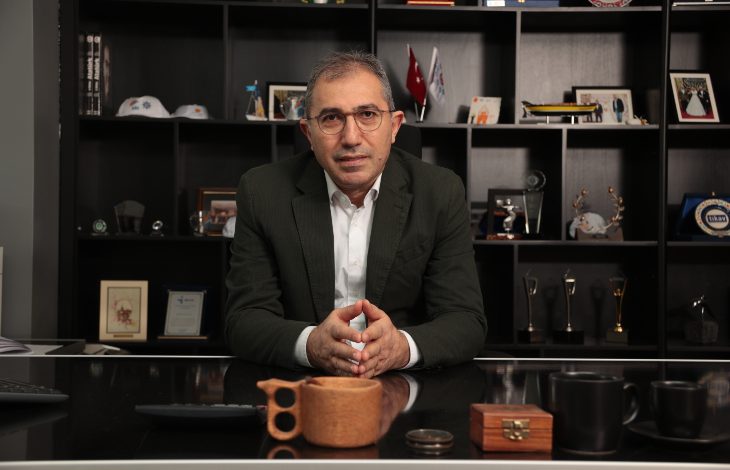 Fotoğrafta bir ofiste, İDO Genel Müdürü Dr. Murat Orhan, takım elbiseli, gözlüklü bir takarak başında oturuyor. Arkasında ödüller ve kurumsal objelerle dolu raflar var. Masada ahşap bir fincan, kupalar ve küçük eşyalar duruyor. Genel hava profesyonel, ciddi ve düzenli.