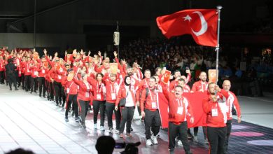 ,Kırmızı–beyaz takım eşofmanları giymiş işitme engelli sporcu grubu, ellerinde Türk bayrağı ve küçük bayrakçıklarla coşkuyla yürüyerek bir açılış töreninde geçiş yapıyor.