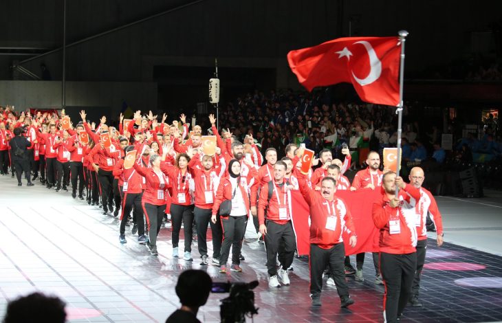 ,Kırmızı–beyaz takım eşofmanları giymiş işitme engelli sporcu grubu, ellerinde Türk bayrağı ve küçük bayrakçıklarla coşkuyla yürüyerek bir açılış töreninde geçiş yapıyor.