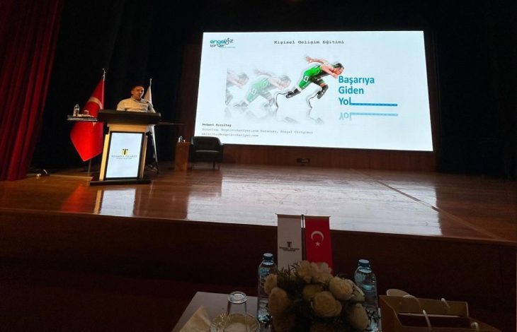 Sahne loş ışıklarla aydınlanmış, ağır ve saygın bir ortam var. Büyük bir projeksiyon perdesinde “Başarıya Giden Yol” başlıklı bir sunum görünüyor. Sol tarafta kürsünün arkasında bir kişi konuşma yapıyor; yanında Türk bayrağı ve İstanbul Ticaret Üniversitesi’nin flaması duruyor. Sahnenin önünde su şişeleri, çiçek aranjmanı ve küçük bayrakların bulunduğu bir masa dikkat çekiyor. Genel atmosfer, profesyonel ve akademik bir eğitim etkinliğini yansıtıyor.