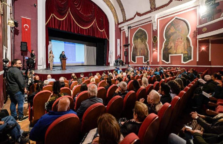 Tarihi bir tiyatro salonunda, kırmızı koltuklara oturmuş kalabalık bir izleyici grubunun karşısında, sahnede bir sunucu ve yanında işaret diliyle çeviri yapan bir kişiyle bir sunum gerçekleştiriliyor.