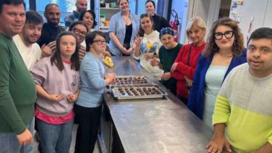 Görselde, bir mutfakta tezgâhın etrafında toplanmış bir grup kişi görülüyor. Tezgâh üzerinde tepsiyle hazırlanmış çikolata ya da kurabiye benzeri yiyecekler var. Katılımcılar gülümseyerek poz veriyor. Ortam, mutfak becerileri eğitimi ya da atölye çalışması havasında.