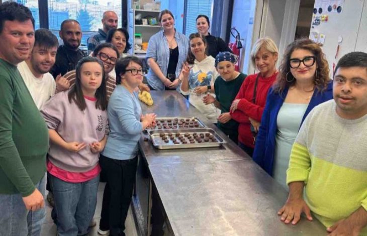 Görselde, bir mutfakta tezgâhın etrafında toplanmış bir grup kişi görülüyor. Tezgâh üzerinde tepsiyle hazırlanmış çikolata ya da kurabiye benzeri yiyecekler var. Katılımcılar gülümseyerek poz veriyor. Ortam, mutfak becerileri eğitimi ya da atölye çalışması havasında.