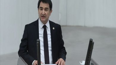 Görselde bir kürsüde konuşma yapan takım elbiseli MHP Genel Başkan Yardımcısı ve İstanbul Milletvekili İsmail Faruk Aksu, görülüyor. TBMM Genel Kurulu’nu andıran geniş, açık renkli bir salonda duruyor. Önünde mikrofonlar, belgeler ve elektronik oylama panelleri var. Ciddi ve resmî bir konuşma anını yansıtıyor.