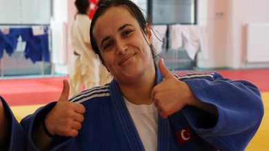 Görselde, mavi judogi giymiş bir kadın sporcu kameraya gülümseyerek iki başparmağını yukarı kaldırıyor. Arka planda antrenman salonu ve diğer judogiler görünüyor. Sporcu enerjik ve gururlu bir ifade taşıyor.