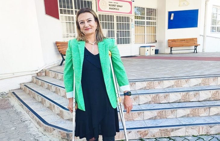 Nusret Şenay Anaokulu binası önünde, yeşil ceketli ve koltuk değnekleriyle ayakta duran öğretmen Hilal Demir’in fotoğrafı.