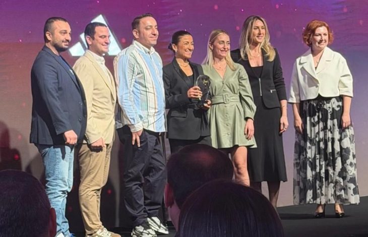 Bir sahnede yan yana dizilmiş yedi kişilik bir grup görülüyor. Hepsi şık ve modern kıyafetlerle poz veriyor. Ortadaki iki kişi küçük, cam görünümlü bir ödül objesini birlikte tutuyor. Arka planda mor ve pembe tonlarda ışıklı bir sahne efekti var. Ön planda ise izleyici siluetleri seçiliyor. Tören atmosferi belirgin.