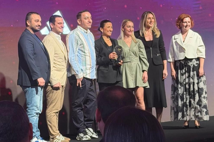 Bir sahnede yan yana dizilmiş yedi kişilik bir grup görülüyor. Hepsi şık ve modern kıyafetlerle poz veriyor. Ortadaki iki kişi küçük, cam görünümlü bir ödül objesini birlikte tutuyor. Arka planda mor ve pembe tonlarda ışıklı bir sahne efekti var. Ön planda ise izleyici siluetleri seçiliyor. Tören atmosferi belirgin.