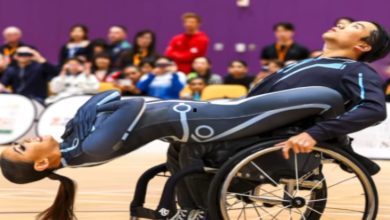 Bir spor salonunda, tekerlekli sandalyedeki bir dansçı partneriyle birlikte etkileyici bir dans figürü sergiliyor. Arkada izleyici topluluğu sahneyi dikkatle izliyor.