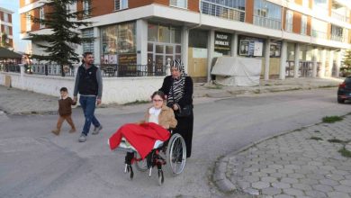 Görselde, tekerlekli sandalyede oturan küçük bir kız çocuğu bulunuyor. Üstü kırmızı bir battaniyeyle örtülü. Sandalyeyi başörtülü bir kadın itiyor. Arkada, bir erkek ve yanında yürüyen küçük bir çocuk yer alıyor. Hepsi bir apartman ve iş yerlerinin bulunduğu bir sokakta ilerliyor.
