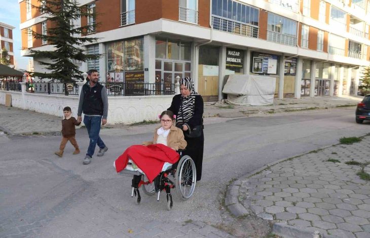 Görselde, tekerlekli sandalyede oturan küçük bir kız çocuğu bulunuyor. Üstü kırmızı bir battaniyeyle örtülü. Sandalyeyi başörtülü bir kadın itiyor. Arkada, bir erkek ve yanında yürüyen küçük bir çocuk yer alıyor. Hepsi bir apartman ve iş yerlerinin bulunduğu bir sokakta ilerliyor.