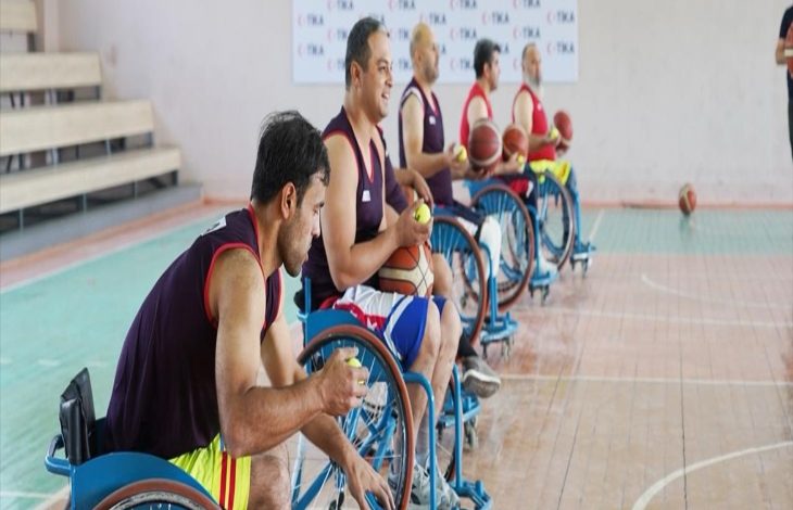 Bir spor salonunda, tekerlekli sandalyede oturan erkek sporcular basketbol toplarıyla antrenman yapıyor. Oyuncular sıralı şekilde dizilmiş ve odaklanmış görünüyor.