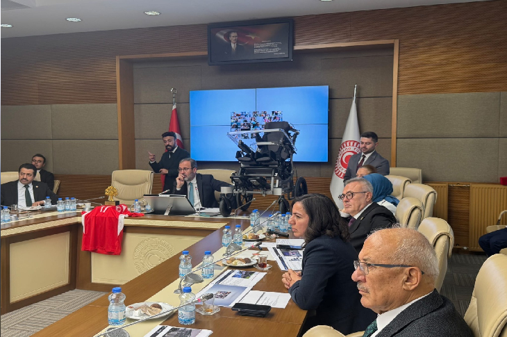 Toplantı salonunda geniş bir masa etrafında toplanmış kişiler görünüyor. Ortada büyük bir kamera düzeneği, arkada iki bayrak ve duvarda büyük ekranlar var. Masada su şişeleri, evraklar ve ikramlar duruyor; ortam resmen yoğun bir komisyon toplantısının nabzını taşıyor.