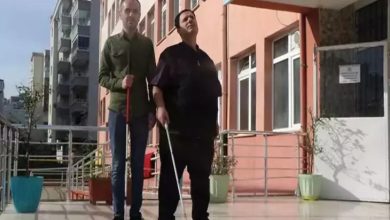 Fotoğrafta, bir okul binasının girişinde iki görme engelli öğretmen yan yana duruyor. Her ikisi de beyaz bastonlarıyla ayakta ve kendinden emin bir duruş sergiliyor. Soldaki öğretmen ince yapılı, yeşil bir gömlek giymiş; sağdaki öğretmen ise daha iri yapılı ve koyu renk bir kıyafet tercih etmiş. Arka planda okulun pembe tonlarındaki dış cephesi, pencereler ve giriş kapısı görünüyor. Güneşli bir hava, zemine düşen ışık ve gölgelerle belli oluyor. Sahne genel olarak güçlü, ilham veren bir birliktelik hissi taşıyor.