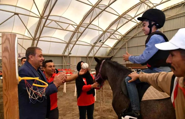 Kapalı manejde düzenlenen etkinlikte, bir çocuk at üzerinde terapi yaparken çevresindekiler onu destekleyip alkışlıyor. Çocuk kask takmış, gülümseyerek topa uzanıyor.