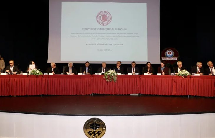 Uzun bir masanın arkasında oturan kalabalık bir grup insan görülüyor. Masanın üzerinde kırmızı masa örtüsü, çiçek aranjmanları ve mikrofonlar bulunuyor. Arkadaki büyük ekranda “Türkiye Büyük Millet Meclisi Başkanlığı” başlıklı bir sunum yansıtılmış.