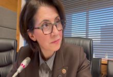 Dr. Aylin Yaman mikrofonun önünde oturuyor ve dikkatle bir yöne bakıyor. Arka planda ofis ortamı ve büyük pencereler görünüyor.