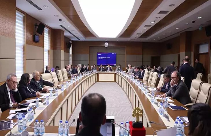 Geniş, modern bir toplantı salonunda uzun U şeklindeki masanın etrafını dolduran çok sayıda kişi görülüyor. Katılımcılar belgelerine eğilmiş, not alıyor ya da konuşmaları takip ediyor. Masaların üzerinde su şişeleri, dosyalar ve mikrofonlar yer alıyor. Salonun ortasında büyük bir ekran bulunuyor. Ortam, yoğun gündemli ve resmi bir komisyon toplantısı atmosferi taşıyor.