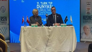 Masada iki kişi yan yana oturmuş, basın toplantısı düzenliyor gibi görünüyor. Arkalarında mavi bir ekran ve “11. Türk Tıp Dünyası Kurultayı” yazısı var. Masada küçük Türk bayrakları, su şişeleri ve mikrofonlar duruyor. Yanlarda SMA tedavisiyle ilgili afişler bulunuyor.