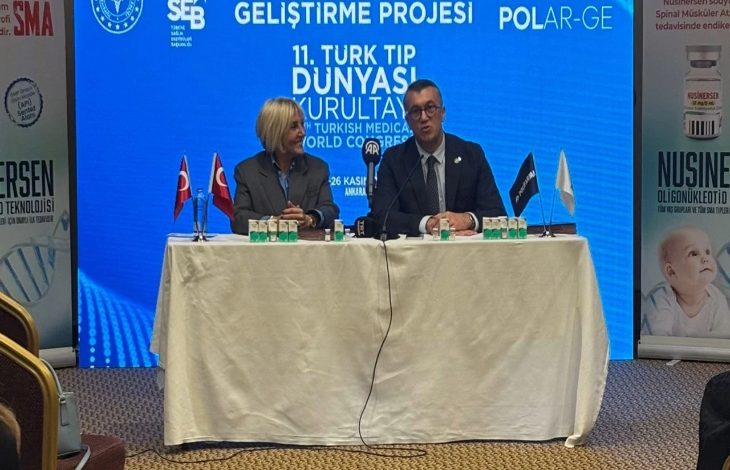 Masada iki kişi yan yana oturmuş, basın toplantısı düzenliyor gibi görünüyor. Arkalarında mavi bir ekran ve “11. Türk Tıp Dünyası Kurultayı” yazısı var. Masada küçük Türk bayrakları, su şişeleri ve mikrofonlar duruyor. Yanlarda SMA tedavisiyle ilgili afişler bulunuyor.