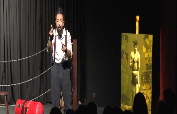 Sahnede tek başına duran bir oyuncu, işaret diliyle performans seriliyor gibi görünüyor. Arka planda boks ringini andıran halatlar, yerde kırmızı bir boks kum torbası ve yanında eski bir boksörün fotoğraflarının yer aldığı sarı tonlu bir pano bulunuyor. Seyirciler karanlıkta, ön planda siluet olarak seçiliyor.