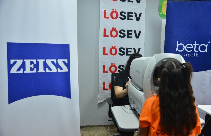 Görselde bir göz tarama etkinliği görülüyor. Sağ tarafta turuncu tişört giymiş bir çocuk, bir göz ölçüm cihazına bakıyor. Cihazın karşısında oturan görevli, ölçümü gerçekleştiriyor. Arka planda LÖSEV’in dikey afişi, yanında ZEISS ve Beta Optik logoları yer alıyor. Etkinlik ortamı oldukça düzenli; sağlık taramasına odaklanan, çocukların görme sağlığını destekleyen bir çalışma yürütüldüğü hissi veriyor.
