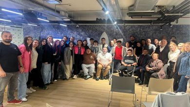 Görselde, geniş bir grup bir araya gelmiş şekilde poz veriyor. Farklı yaş ve özelliklerde birçok kişi ayakta veya sandalyelerde oturarak gülümsüyor. Ortam modern bir iç mekân; tavan aydınlatmaları, havalandırma boruları ve ahşap zemin dikkat çekiyor. Genel atmosfer sıcak, kapsayıcı ve topluluk ruhunu yansıtan bir buluşma izlenimi veriyor.