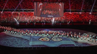 Büyük bir stadyumda gerçekleştirilen görkemli bir tören görülüyor; sahnenin ortasında ışık gösterileri ve semboller yer alırken, sahada Türk bayrakları taşıyan sporcular yürüyüş yapıyor ve tribünlerde kırmızı ışıklarla aydınlatılmış kalabalık bir izleyici topluluğu bulunuyor.