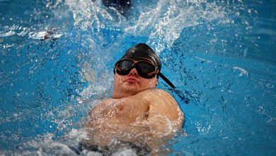 Bir yüzme havuzunda, bone ve gözlük takmış bir sporcunun sırtüstü yüzme sırasında suyu güçlü şekilde yararak ilerlediği an görülüyor. Su sıçramaları ve sporcunun odaklanmış ifadesi dikkat çekiyor.