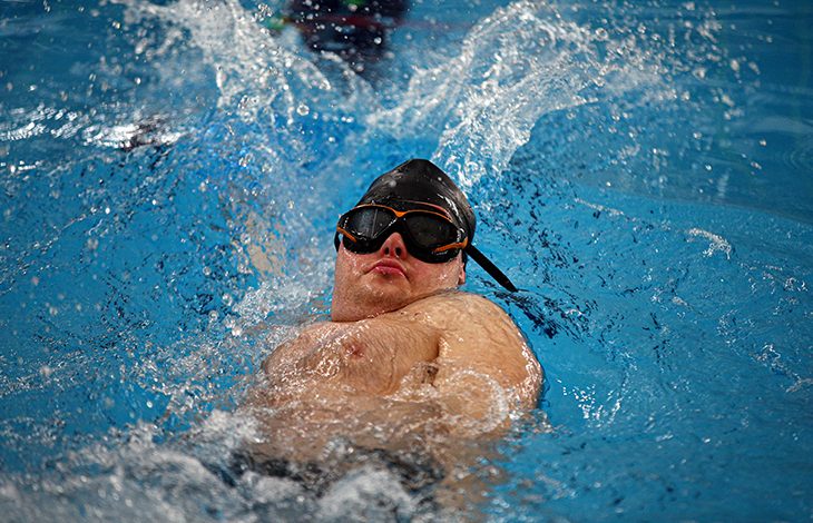 Bir yüzme havuzunda, bone ve gözlük takmış bir sporcunun sırtüstü yüzme sırasında suyu güçlü şekilde yararak ilerlediği an görülüyor. Su sıçramaları ve sporcunun odaklanmış ifadesi dikkat çekiyor.