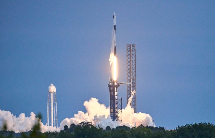 Açık havada bir fırlatma rampasında roketin kalkış anı görülüyor. Roketin altından yoğun alev ve beyaz duman yükselirken, arka planda metal kuleler ve açık mavi gökyüzü yer alıyor. Sahne güçlü, tarihi bir uzay yolculuğunun başlangıç anını yansıtıyor.