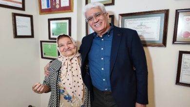 Duvarı sertifika ve plaketlerle dolu bir odada, yaşlı bir kadın ile yanında duran bir adam yan yana gülümseyerek poz veriyor. Kadın başörtülü, çiçek desenli elbise ve hırka giyiyor; adam ise koyu renk bir ceket ve gömlekle şık bir görünüme sahip.