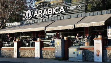 Cadde üzerinde yer alan Arabica Coffee House şubesi görülüyor. Geniş tenteler altındaki açık oturma alanında müşteriler masalarda oturuyor. Üstte büyük “Arabica Maestro Coffee House” tabelası var; mekân canlı, modern ve davetkâr bir kafe atmosferi sunuyor.