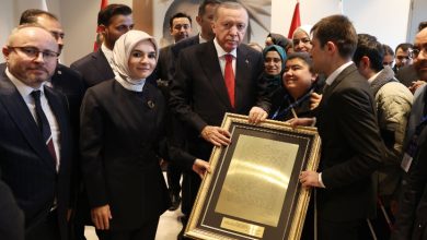 Görselde kalabalık bir grup insan bir arada duruyor. Ortada takım elbiseli bir erkek, büyük boyutlu çerçeveli bir tabloyu iki yanındaki kişilerle birlikte tutuyor. Çerçevenin içinde altın tonlarında bir metin yer alıyor. Sol tarafta başörtülü bir kadın, sağ tarafta ise genç bir erkek bulunuyor. Arkada farklı yaş ve görünümde birçok kişi görüntüye dahil olmuş. Ortam resmi ve tören havasında, arka planda bayraklar görülüyor.