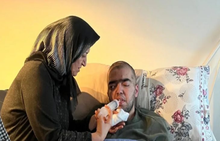 Tayyip Ömer Uzuner yatarken, yaşlı kadın şırınga benzeri bir aparatla ağızdan besin vermeye çalışılıyor.