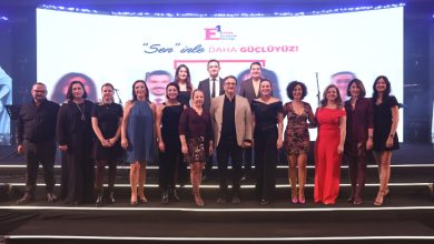 Bir sahnede yan yana dizilmiş, şık kıyafetler giymiş kalabalık bir grup, arkada etkinliğin logosu ve sloganının yer aldığı büyük bir ekran önünde kameraya poz veriyor; ortam resmi ve kutlama havasında.