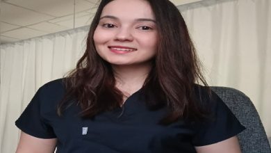 Bir muayenehane ya da klinik ortamını andıran perdeli bir arka planda, sağlık çalışanı kıyafeti giymiş genç bir kadın kameraya doğru gülümseyerek oturuyor. Doğal ışıkla aydınlanan sade ve profesyonel bir görüntü.