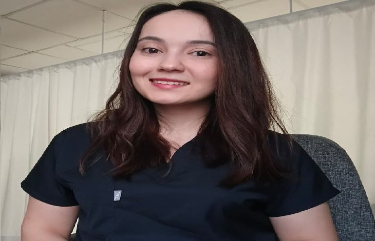 Bir muayenehane ya da klinik ortamını andıran perdeli bir arka planda, sağlık çalışanı kıyafeti giymiş genç bir kadın kameraya doğru gülümseyerek oturuyor. Doğal ışıkla aydınlanan sade ve profesyonel bir görüntü.