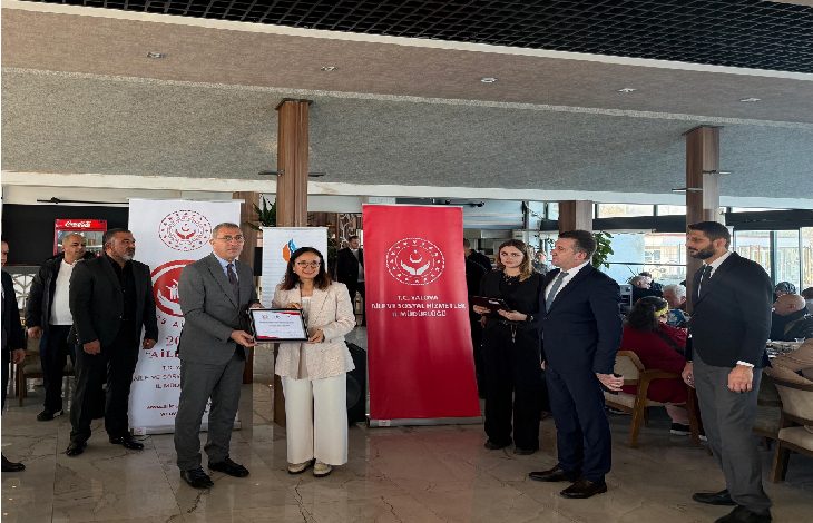 Fotoğrafta bir tören anı görülüyor. Ortada, bir kadın ile bir erkek birlikte bir sertifika tutarak kameraya poz veriyor. Arkalarında Aile ve Sosyal Hizmetler Bakanlığı logolu iki büyük pankart bulunuyor. Sağda ve solda takım elbiseli yetkililer ile davetliler ayakta duruyor. Mekân ferah, gün ışığı alan bir salon ve arkada oturan kalabalık bir topluluk dikkat çekiyor.
