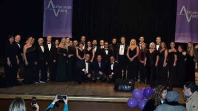 Bir sahnede, şık siyah kıyafetler giymiş kalabalık bir koro ve katılımcılar toplu halde poz veriyor; arkada Alzheimer Vakfı afişleri, önde ise izleyiciler fotoğraf çekiyor.