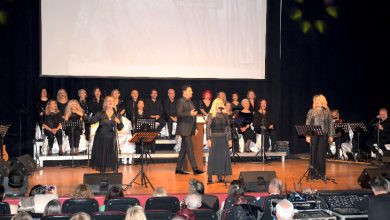 Bir konser salonunda sahnede koro ve solistlerin yer aldığı, arkada müzisyenlerin bulunduğu kalabalık bir müzik dinletisi görülüyor. Ön planda seyirciler otururken, sahnede toplu performans ve canlı bir etkinlik atmosferi dikkat çekiyor.