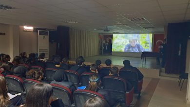 Bir konferans salonunda sıralara oturmuş öğrenciler, perdede yansıtılan bir belgeseli dikkatle izliyor. Işıklar kısık, sahnede sunum alanı ve Türk bayrağı var. Ortam, farkındalık ve empati odaklı bir etkinliğin sessiz ama yoğun havasını taşıyor.