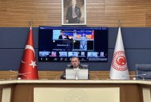 Görselde, TBMM’de bir toplantı salonu görülüyor. Ortada kürsüde oturan bir kişi bilgisayar başından çevrim içi toplantıyı yönetiyor. Arka planda büyük ekranda çok sayıda katılımcının yer aldığı video konferans görüntüsü bulunuyor; salonun iki yanında Türk bayrağı ve TBMM bayrağı yer alıyor.