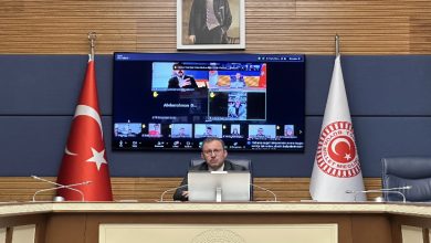 Görselde, TBMM’de bir toplantı salonu görülüyor. Ortada kürsüde oturan bir kişi bilgisayar başından çevrim içi toplantıyı yönetiyor. Arka planda büyük ekranda çok sayıda katılımcının yer aldığı video konferans görüntüsü bulunuyor; salonun iki yanında Türk bayrağı ve TBMM bayrağı yer alıyor.