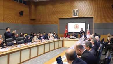 Türkiye Büyük Millet Meclisi’nde, uzun bir toplantı masası etrafında çok sayıda katılımcının yer aldığı resmi bir komisyon toplantısı görülüyor. Salonda Türk bayrakları, kürsü ve arka planda TBMM amblemi bulunurken, katılımcılar not alıyor ve konuşmacıyı dikkatle dinliyor.