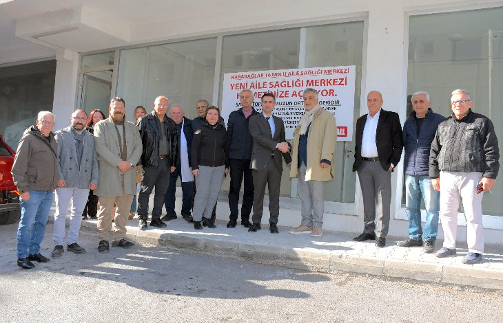 Bir aile sağlığı merkezinin önünde, gündüz ışığında yan yana dizilmiş kalabalık bir grubun toplu fotoğrafı görülüyor. Arka planda sağlık merkezi camı ve açılışa dair bilgilendirme yazısı yer alırken, katılımcılar resmi ve sade bir şekilde kameraya bakıyor.